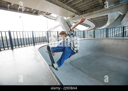 Ritratto laterale del giovane uomo che guida lo skateboard e fa trucchi sulla rampa allo skatepark all'aperto Foto Stock