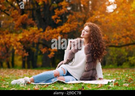 Bella ragazza snella in un gilet di pelliccia sta riposando in autunno nel parco e seduto sul prato sul prato Foto Stock