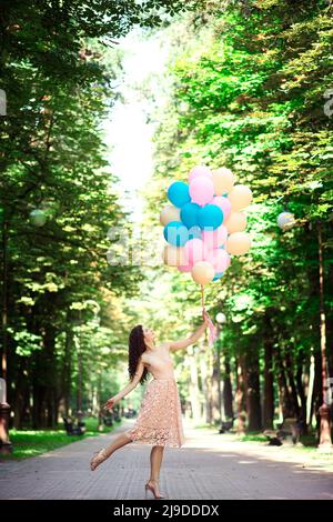 Ragazza slanciata bella alta in un abito e capelli ricci sta tenendo palloncini multicolore nel parco in estate Foto Stock