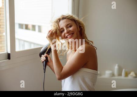 Ragazza allegra e graziosa che asciuga i capelli con l'asciugacapelli e sorride in bagno al mattino. Foto Stock
