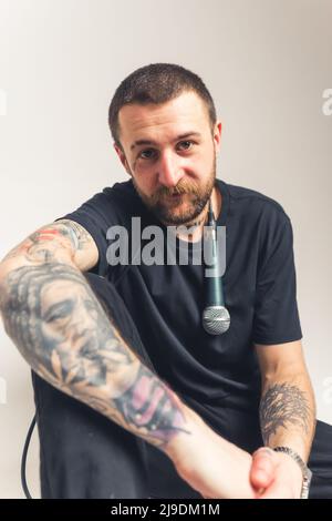 In studio verticale è stato girato un creatore di musica da tatuaggio di mezza età in una t-shirt nera con un microfono che pende sulla spalla guardando la fotocamera. Foto di alta qualità Foto Stock