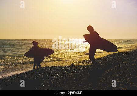 Silhouette Padre e Figlio con Surfboard a Beach Against Sky durante il tramonto. Retroilluminazione durante il tramonto con cielo e riflessi spettacolari. Foto Stock