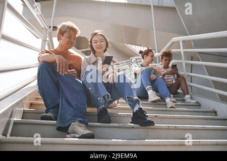 Riprese a tutta lunghezza illuminate da sole di adolescenti diversi utilizzando smartphone all'aperto mentre si siede su scale di metallo Foto Stock