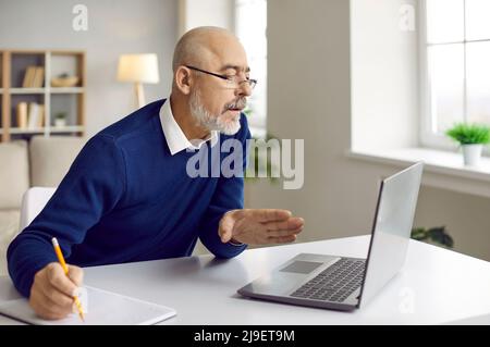 Uomo anziano seduto alla scrivania con notebook, che guarda il webinar e prende appunti su carta Foto Stock