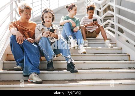 Arioso scatto a tutta lunghezza di adolescenti diversi utilizzando smartphone all'aperto mentre si siede su scale di metallo alla luce del sole Foto Stock