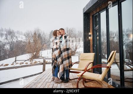 Intera lunghezza di uomo e donna avvolto in coperta in piedi vicino sedie fuori casa di legno con finestre panoramiche. Coppia felice abbraccia all'aperto sotto la neve d'inverno. Foto Stock