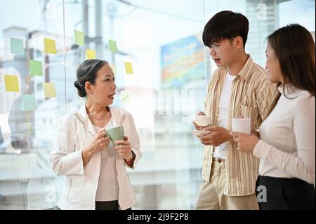 Il capo asiatico senior di successo parla e condivide l'idea creativa con due giovani dipendenti in ufficio durante la pausa caffè. Foto Stock