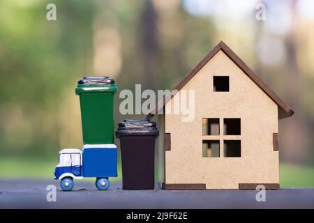 Carrello per rifiuti giocattolo con bidoni per rifiuti in piedi da un piccolo modello di casa in legno su uno sfondo di foresta. Raccolta di rifiuti in comunità rurali. Foto Stock