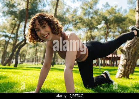 Felice giovane bella donna rossa indossando abiti sportivi facendo esercizi di cane da uccello e guardando la macchina fotografica con grandi sorrisi sul verde parco della città, outd Foto Stock