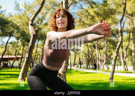 Bella donna rossa che indossa abiti sportivi facendo squat e guardando via sul verde parco della città, all'aperto. Vita sana, buona forma del corpo e all'aperto Foto Stock