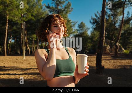Felice giovane donna rossa che indossa abiti sportivi in piedi sul parco della città, all'aperto con tazza da caffè da asporto e parlare al telefono con gli amici mentre l Foto Stock