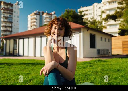 Giovane donna rossa sportiva che indossa un reggiseno sportivo e pantaloni yoga blu sul parco cittadino, all'aperto seduto su un tappeto yoga sorridendo e posando o guardando la ca Foto Stock