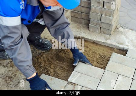 Il muratore in abiti da lavoro si siede sul marciapiede e stende le lastre di pavimentazione. Vista dell'uomo in attività all'aria aperta. Costruttore professionista fa organizzazione del territorio giorno d'estate. Scena reale. Foto Stock