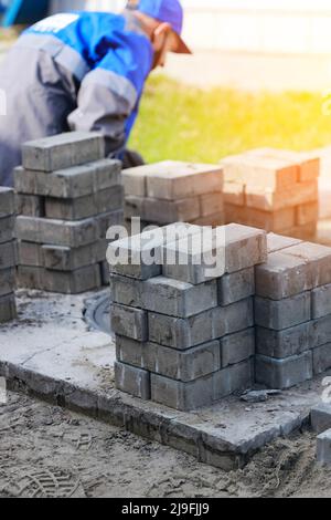 Il muratore in abiti da lavoro si siede sul marciapiede e stende le lastre di pavimentazione. Vista dell'uomo in attività all'aria aperta. Costruttore professionista fa organizzazione del territorio giorno d'estate. Scena reale. Foto Stock