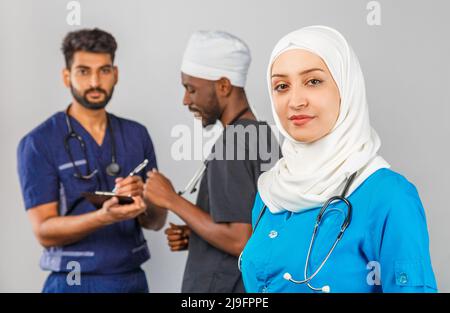 Gruppo di paramedici sorridenti su sfondo grigio. donna medico musulmana con colleghi chirurgo africano e indiano Foto Stock