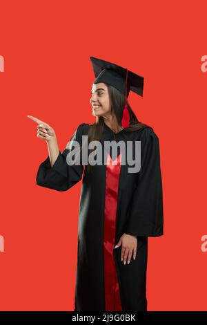 Vista laterale di bella ragazza in piedi, sorridente, mostrando da finger.Beautiful giovane donna che si laurea dal college, indossando abito laureato e Mortarboard. Isolato su sfondo rosso. Foto Stock