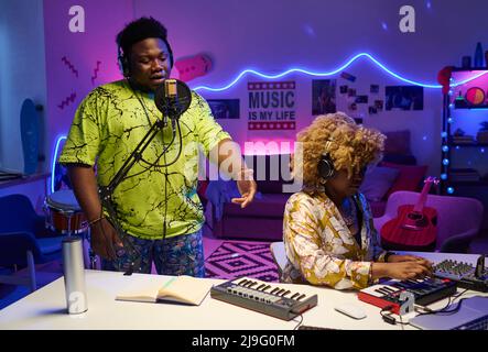 Elegante uomo afro-americano gen Z che suona il microfono mentre la sua ragazza suona il sintetizzatore e regola il suono sulla console di mixaggio Foto Stock