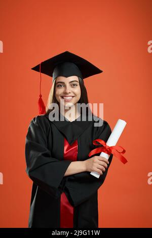Vista frontale della bella ragazza che si laurea da college, università, scuola superiore. Bruna giovane donna in piedi con mani incrociate, con diploma, sorridente. Concetto di gioventù. Foto Stock