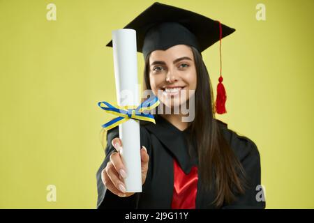 Vista frontale della ragazza brunetta che mostra diploma ucraino. Bella donna in piedi, guardando la macchina fotografica, sorridente, indossando la tavola da mortarboard e abito laureato. Isolato su sfondo verde. Foto Stock