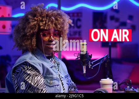 Orizzontale medio primo piano di gioiosa giovane afroamericana indossando abiti alla moda seduti in studio di trasmissione sorridendo alla macchina fotografica Foto Stock
