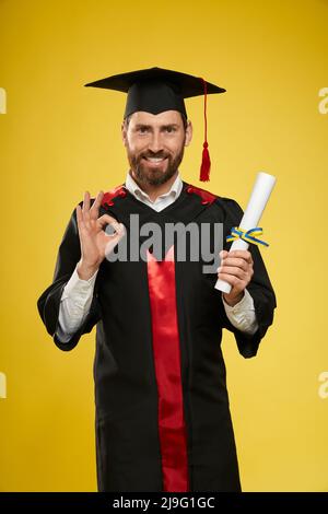 Vista frontale dello studente che indossa abito e mazzetto con diploma, mostrando Va bene. Giovane che si laurea al college, felice, sorridente. Concetto di istruzione e gioventù. Foto Stock