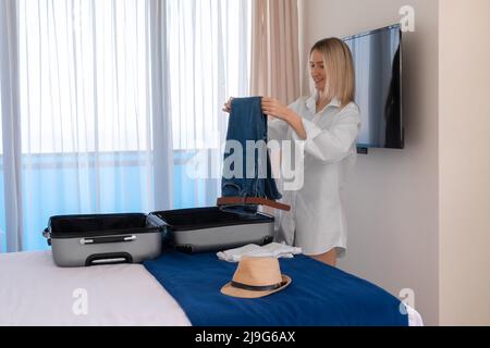 Allegra giovane donna felice in una camicia bianca raccoglie o scompatta le cose da una valigia che si trova sul letto. Concetto di viaggio, vacanza Foto Stock