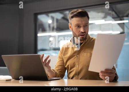 Scioccato uomo d'affari che legge i documenti che hanno problema usando il laptop in ufficio Foto Stock