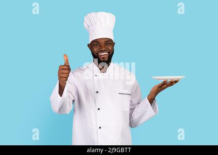 Bel cuoco nero in uniforme tenendo piatto vuoto e mostrando Thumb Up Foto Stock