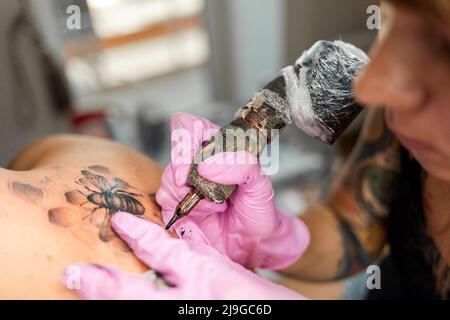 Il primo piano di macchina e mani in guanti di lattice rosa rende un tatuaggio design alla schiena del cliente. Concetto di tatuaggio del mondo giorno. Foto Stock