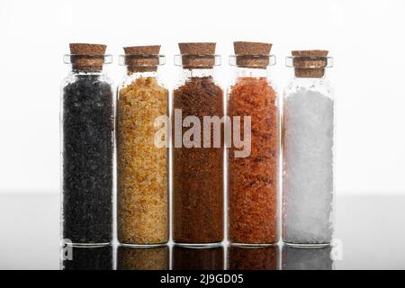 Concetto di cibo. Varie spezie in bottiglie di vetro a forma di cilindro con sughero su fondo bianco su superficie riflettente scura. Sale variopinto Foto Stock