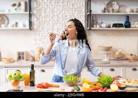 Giovane donna che mangia insalata a tavola con verdure biologiche, gustando una dieta sana, in piedi in cucina interna leggera Foto Stock