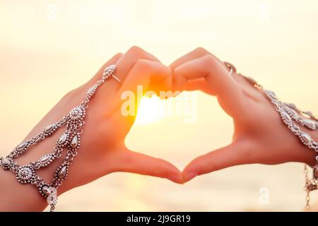 Le mani femminili in forma di cuore contro il cielo passano raggi di sole. Mani in un lussuoso doppio braccialetto argento gioielli e anello stile orientale a forma di Foto Stock