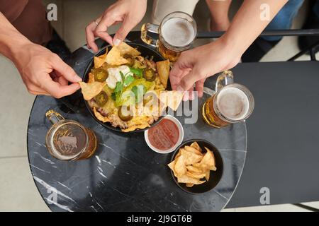 Mani di persone che bevono birre con ciotola di nacho patatine con riso, fette di jalapeno, guacamole e panna acida, vista dall'alto Foto Stock