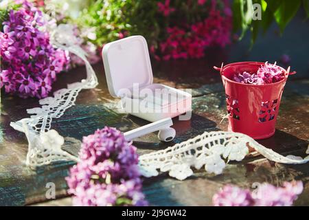 Stile di vita estetico piatto, cuffie wireless, fiori lilla e decorazioni in pizzo. Ascoltare musica o audiolibro, partecipare a webinar o chat online, parlare al telefono in giardino soleggiato Foto Stock