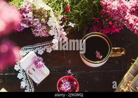 Stile di vita estetico piatto, cuffie senza fili, fiori lilla decorazioni e tazza di caffè. Ascolto di musica o audiolibro, webinar o chat online, conversazione telefonica in giardino Foto Stock