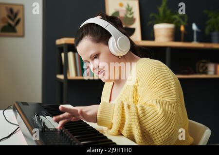 Moderna giovane donna caucasica con sindrome di Down indossando abiti casual e cuffie che regolano le impostazioni audio sulla tastiera elettronica Foto Stock