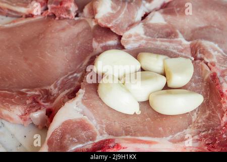 Un primo piano di una manciata di spicchi d'aglio sopra alcune braciole di maiale crudo con sale su un tagliere. Preparazione della carne e macelleria. Foto Stock