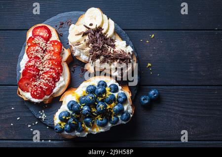 Toast vegetariani fruttati per colazione su sfondo di legno scuro, vista dall'alto. Fette di pane con ricotta, bacche, banana, cioccolato e semi. Foto Stock