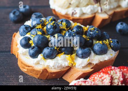 Toast Berry vegetariani per colazione su sfondo di legno scuro, primo piano. Fette di pane con ricotta, bacche, banana, cioccolato e semi. Foto Stock