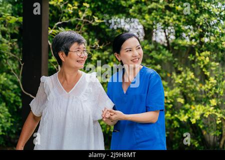 Donna felice caregiver in scrub blu che tiene la mano della donna anziana e guardando via con il sorriso mentre cammina vicino alberi lussureggianti in giardino insieme Foto Stock