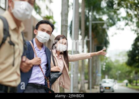 Giovane donna asiatica d'affari in maschera protettiva cattura taxi quando si affretta a incontrare Foto Stock