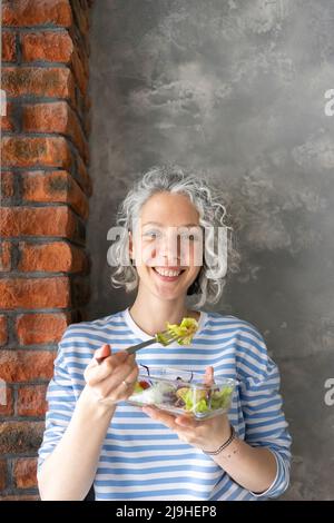 Donna felice che mangia insalata a pranzo di fronte al muro Foto Stock