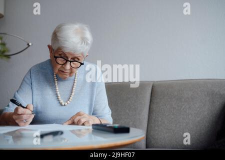 Donna anziana che fa i documenti seduti sul divano a casa Foto Stock