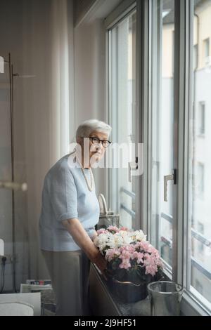 Donna anziana in piedi da fiori e finestra a casa Foto Stock