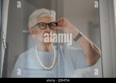 Donna anziana che indossa occhiali guardando attraverso la finestra Foto Stock
