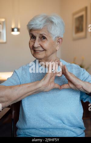 Donna anziana felice che fa segno di cuore con le mani a casa Foto Stock