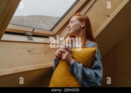 Giovane donna che tiene una tazza di caffè guardando attraverso la finestra Foto Stock