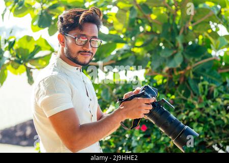 bel fotografo indiano e sicuro con una grande fotocamera professionale che scatta foto scattando foto sulla spiaggia.sessione fotografica in estate Foto Stock