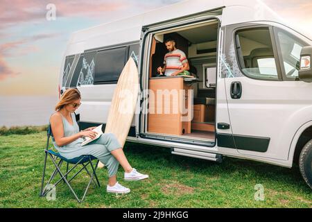 Uomo che cucinano le verdure in un furgone del camper mentre la donna legge all'aperto Foto Stock