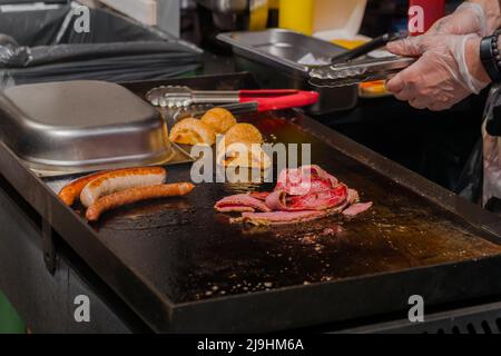 Preparazione di panini con hamburger, fette di pancetta e salsicce lunghe sul grill Foto Stock